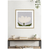 Framed minimalist wall art print of a white tulip with two green leaves against a light gray background, hanging above a wooden console table with books, mug, and decor.