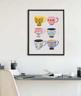 Framed print of six colorful coffee cups arranged in two columns with bright botanical patterns on a white wall above a modern desk.