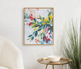 Framed botanical abstract print with jewel tones of green leaves, red and yellow flowers, hanging on a white wall above a round gold table with a white bowl and tall grass in a modern room corner.