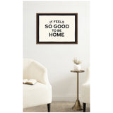 Framed black and white typography wall art reading 'It feels so good to be home' above minimalist seating with off-white textured chairs and a small round brass side table with a white vase.