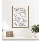 Abstract black continuous line shapes forming interconnected organic patterns on white canvas, framed in natural wood and hung above a white shelf with lamp, photo frame, and potted plant.