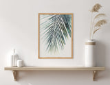 Wood-framed palm leaf print above a beige wooden shelf holding two white vases on the left and a tall white vase with beige pampas grass on the right against a white wall.