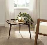 Round dark brown metal coffee table with scalloped edge and slender legs, holding a green trailing plant, two candles, and a book in a sunlit neutral room.