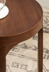 Close-up of a round wooden table with smooth edges and rich brown finish, placed on a beige rug with dark patterned designs.