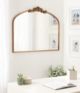Arched mirror with a carved floral crest on top, framed in brown wood, hanging on a white wall above a white desk with plants, a candle, stationery, and an open notebook.