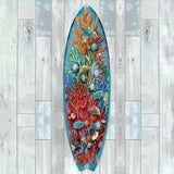 Colorful surfboard-shaped wall art featuring vibrant orange and red coral, sea shells, and blue-green tropical fish on a blue background, displayed on a light wood-paneled wall.