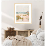 Soft pastel painting of a sandy beach with sea dunes and gentle blue-green waves, framed with a light wooden border, hanging above a neutral-toned bed and cabinet.