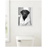 Black Labrador wrapped in a white towel, portrayed in a black and white framed canvas hung above a white toilet against a light wall near a window.
