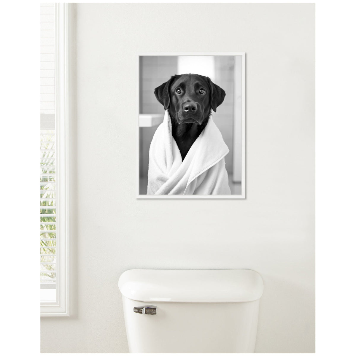 Black Labrador wrapped in a white towel, portrayed in a black and white framed canvas hung above a white toilet against a light wall near a window.