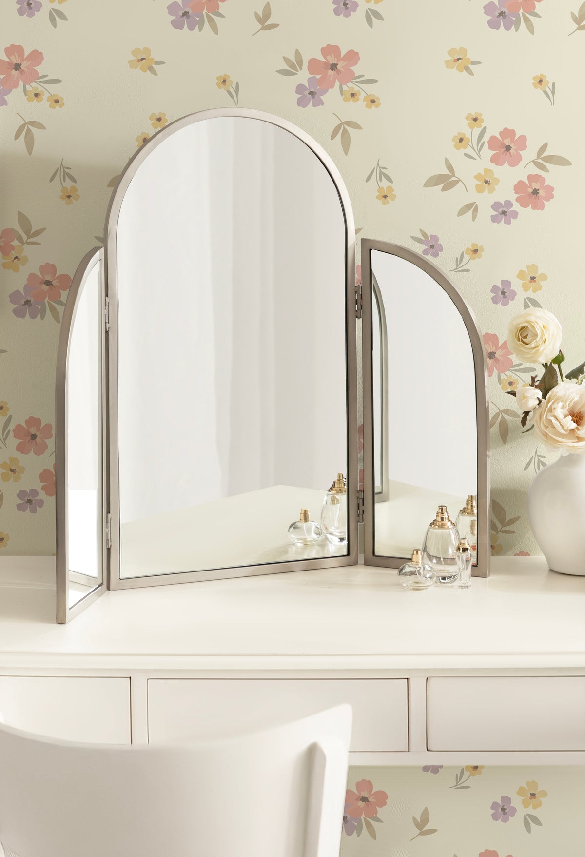 Arch-shaped tri-fold metal mirror with a neutral frame on a white vanity, reflecting a softly lit room with floral wallpaper and a vase of roses nearby.