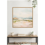 Framed square canvas print of a soft pastel landscape featuring sandy foreground, greenish bushes, and a distant blue ocean under a pale sky, hung above a wooden console with neutral decor.