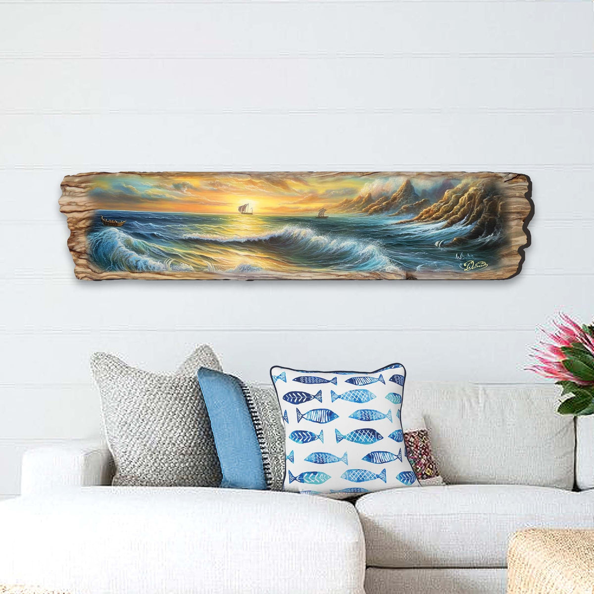 Long wooden panel featuring a painted ocean wave scene with a small sailboat near sunset and dramatic clouds, hung above a white couch with fish-patterned pillows.