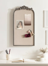 Tall arched pinboard with ornate gray frame holding a photo, paint swatches, and fabric samples, mounted on a light wall above a white desk with stationery and a vase.