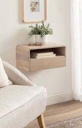 Light wood floating side table with rounded edges mounted on a white wall, featuring an open shelf with books and a top surface holding a small potted plant and glasses.