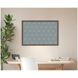 Rectangular pinboard with muted blue fabric featuring small light botanical block print motifs, framed in dark wood, mounted on a beige wall above a light wood desk.