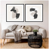 Two black-framed abstract prints with layered neutral and black organic shapes hang above a light gray sofa with a gray throw and round pillow, a black wicker coffee table, and a dog.