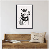 Black and white linocut print of three stylized birds stacked vertically with simple plant motifs, framed and hung above a rustic wooden bench with a "welcome home" pillow.