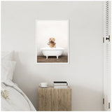 Framed canvas of a golden retriever puppy sitting in a white clawfoot bathtub filled with bubbles, holding a brown rope toy, hung on a white wall above a rustic wooden bedside table.