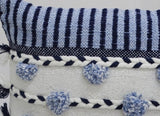 Close-up of a white woven pillow with navy blue braided trim and soft blue pom-poms, topped by a navy and white striped fabric section.