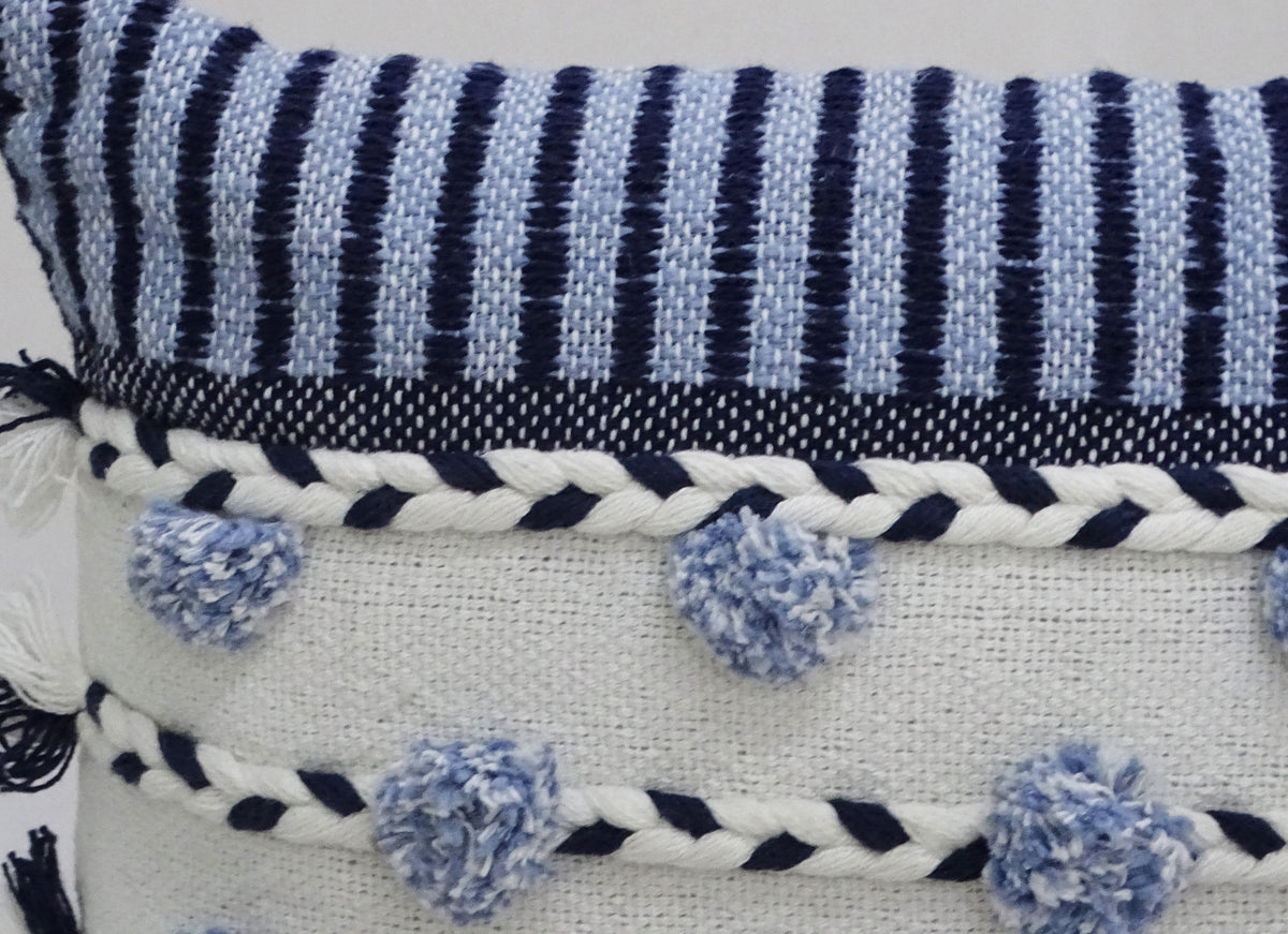 Close-up of a white woven pillow with navy blue braided trim and soft blue pom-poms, topped by a navy and white striped fabric section.