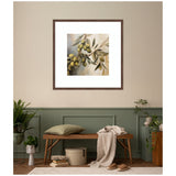 Square framed wall art print depicting an olive tree branch with green olives and leaves in soft, natural tones above a wooden bench with beige cushion and draped cream throw in a minimalist room.