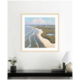 Framed coastal landscape painting depicting sandy shorelines curving alongside blue water and green marshlands under a pastel sky, hanging above a black table with plants and books.