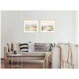 Two framed abstract landscape prints with soft beige, green, and gray tones hung side by side above a beige sofa decorated with patterned black and white pillows and a chunky knit throw.