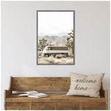 Rustic vintage van parked in a desert landscape with Joshua trees and distant mountains, framed in a muted, natural color palette above a wooden bench with a welcome home pillow.
