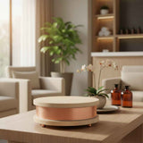 Round wooden container with a copper band around its middle, placed on a wooden coffee table in a softly lit living room with beige chairs and a potted plant in the background.