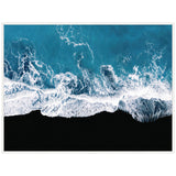 Aerial view of ocean waves crashing onto a stark black sand beach, with deep blue water and white sea foam creating dynamic patterns.