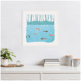 Linocut print of stylized fish swimming in blue water beneath patterned tree trunks, framed in white and hanging on a white wall above a minimalist desk with books and plant.