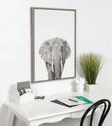 Black and white detailed elephant portrait printed on vertical canvas, framed in light wood, hanging above a white desk with plant and stationery.