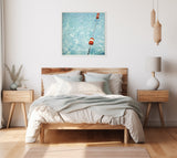 Framed photo above wood bed showing orange and white buoys floating on a rippling blue pool surface, against a clean white wall in a minimalist bedroom.
