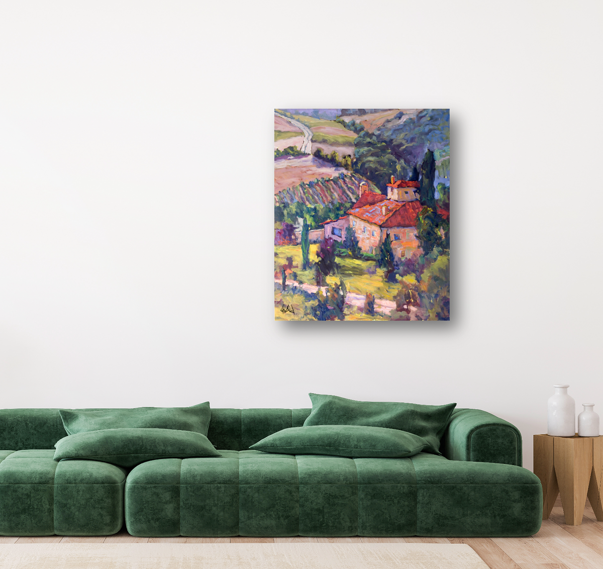 Impressionist-style canvas painting of a vineyard landscape with red-roofed houses, green trees, and rolling hills in vibrant earthy tones hanging on a white wall above a green velvet sofa