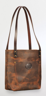 Brown leather tote bag with long double straps, featuring a front pocket embossed with a circular logo and brass rivet details at the strap attachments.