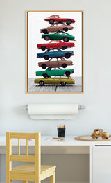 Vertical canvas art depicting a stack of seven vintage cars in various colors, including red, brown, green, and blue, framed in light wood on a white wall.