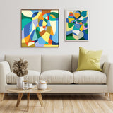 Two abstract colorful paintings above a beige sofa with a green pillow, featuring organic and geometric shapes in warm and cool tones, set against a plain light wall.