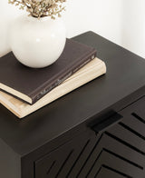 Black floating side table with a slatted, chevron-patterned door, topped with a white ceramic vase holding dried flowers and two stacked hardcover books.