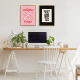 Minimalist workspace with wooden desk and white chair, featuring two framed prints on the wall: pink background with red dice and 'Feeling Lucky' text, and black background with 'Lucky You' card design.