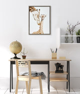 Framed portrait of a mother giraffe nuzzling a baby giraffe, displayed above a wooden desk with a globe, small plant, and elephant figurines.