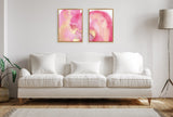 Two framed abstract canvas artworks with pink and gold watercolor textures hang side by side above a white three-seat sofa with four cushions, in a minimalist living room.