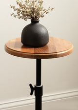 Round wood table top with warm brown tones supported by a black adjustable metal stand, featuring a matte black vase holding delicate dried flowers.