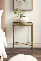 Slim side table with textured brass top and thin bronze metal legs crossed at base, holding a white ribbed vase with green leaves and a pair of glasses in a cozy bedroom corner.