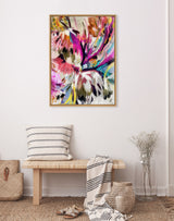 Abstract floral painting with vibrant splashes of pink, purple, yellow, and green framed in light wood, hanging above a wooden bench with striped pillow and draped blanket.
