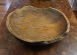 Large round live edge bowl carved from a single teak wood piece, showing smooth interior with natural grain and rough textured outer rim on a parquet wood surface.