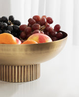 Brushed brass ribbed metal bowl filled with red and black grapes, oranges, and a peach, shown on a white surface with a soft background.