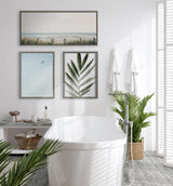 Bright white bathroom featuring a freestanding tub, framed botanical and abstract art on the wall, and lush green plants in woven baskets by shuttered windows.