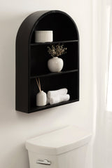 Black matte arch-shaped wall shelf with three shelves displaying a white ribbed jar, a white vase with dried flowers, and towels beside a reed diffuser, mounted above a white toilet.
