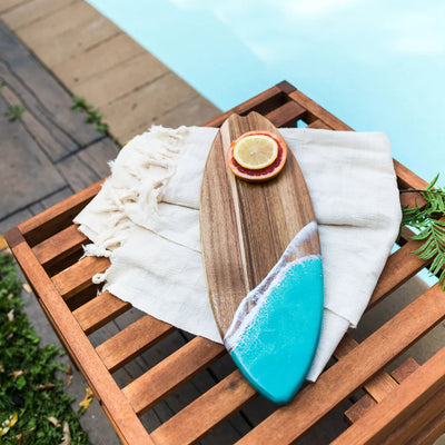 Wooden surfboard-shaped charcuterie board with blue resin accent, holding cheese and fruit on a patio table
