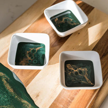 Three small white bowls with green and gold resin designs on a wooden cutting board.